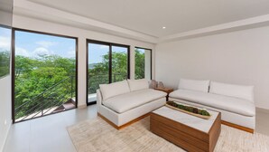 A modern living room with a sectional sofa, a wooden coffee table, and large windows offering a view of greenery and a balcony. - Stunning ocean view house in CocoBay Estates (Coco)
