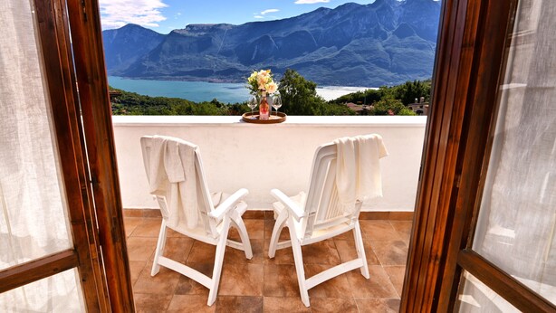 Un balcone con due sedie bianche, un vaso con fiori e una bottiglia. Si può ammirare una vista panoramica su montagne e lago attraverso la porta aperta.