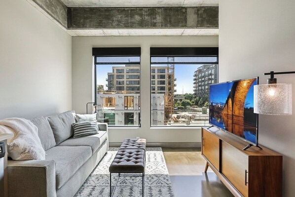 Ein modernes Wohnzimmer mit einem grauen Sofa, einem Holzschrank und einem Fernseher. Große Fenster bieten Blick auf eine Stadtlandschaft und eine Baustelle.