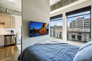 A modern bedroom with a large bed, a cityscape view through large windows, and a kitchen area with wooden cabinets and a dishwasher. - Sable 308 - Alcove (Minneapolis)