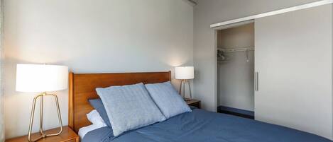 A bedroom with a wooden headboard, two bedside tables with lamps, a large bed with blue bedding, and a walk-in closet with a mirror and clothes rack.