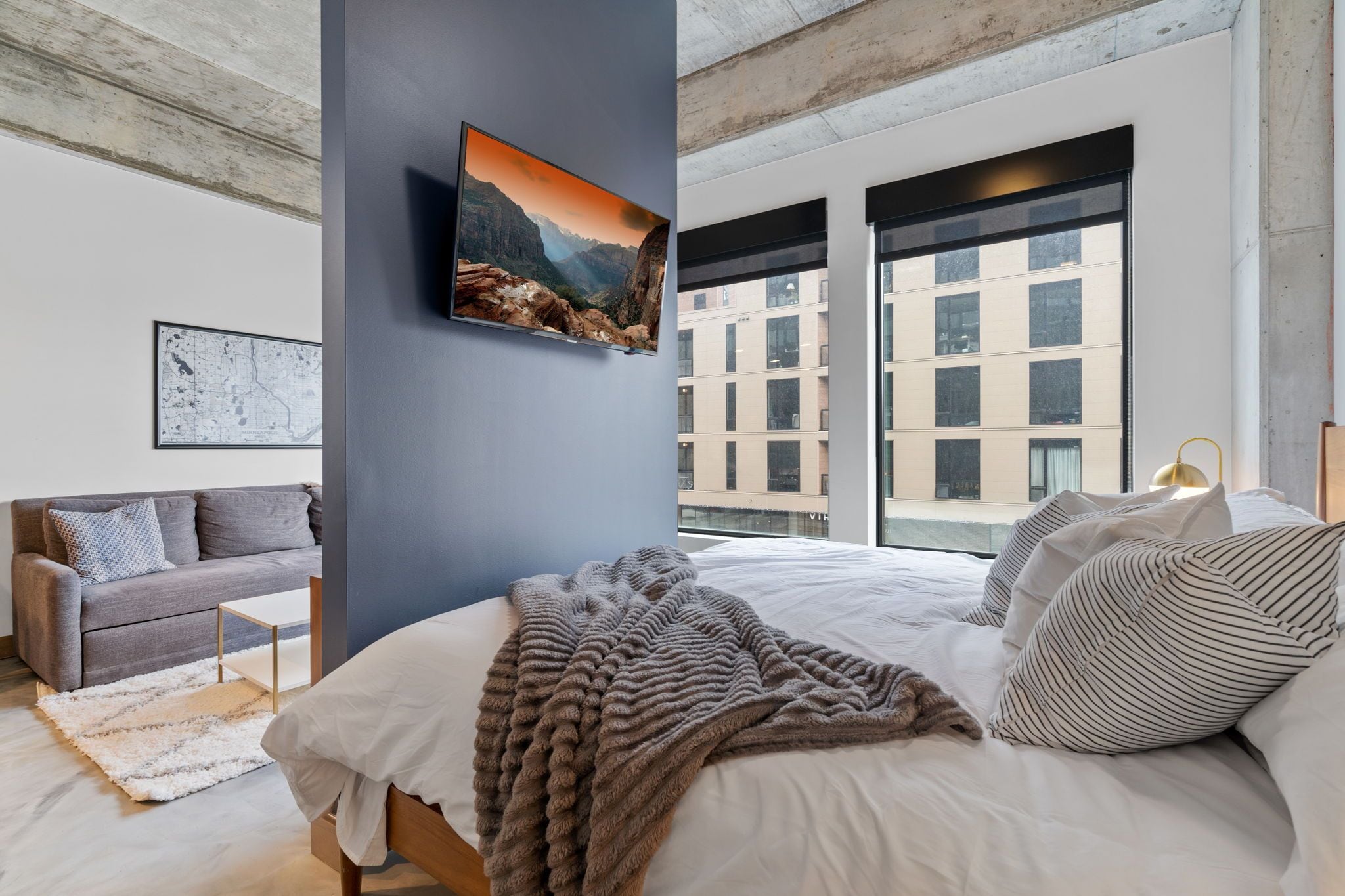 Un dormitorio moderno con una cama grande, un sofá gris, una mesita pequeña y un ventanal con vistas a la ciudad.