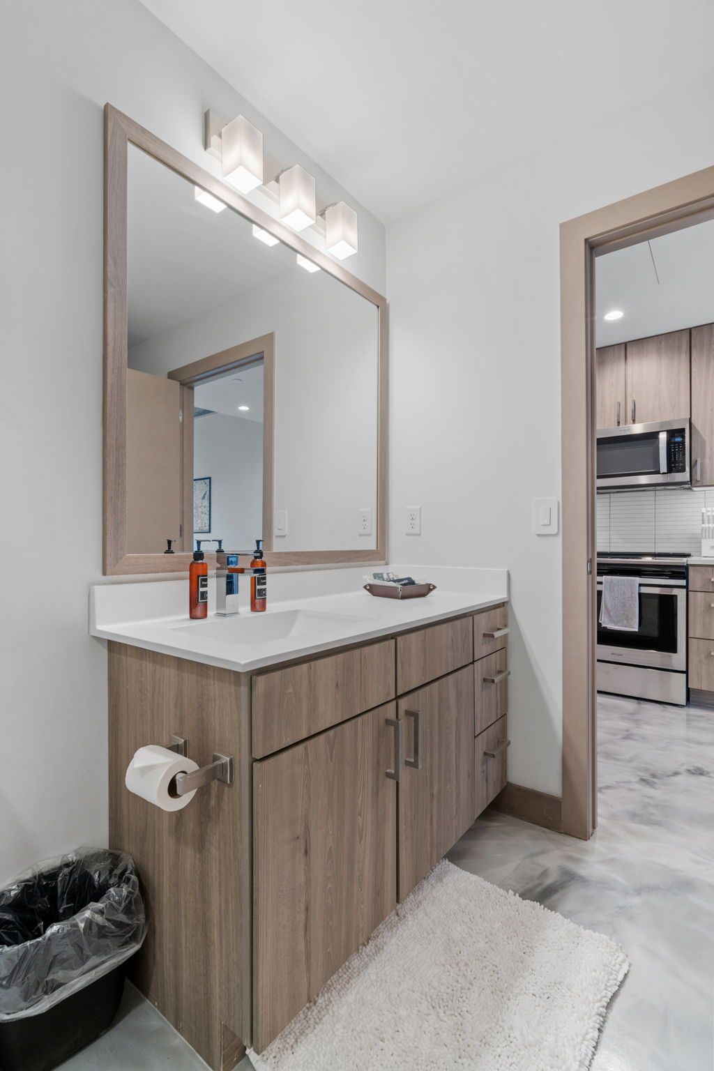 A modern bathroom with a large mirror, wooden cabinets, a white countertop, and a trash bin.