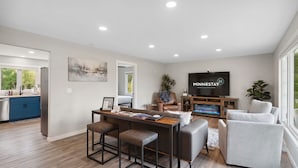A modern living room with a TV, seating, and a kitchen area. - The Cottage at Enchanted Pines (Sauk Rapids)