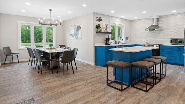 A modern kitchen with a central island, blue cabinets, and a dining area with a chandelier. - The Cottage at Enchanted Pines (Sauk Rapids)