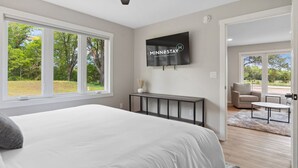 A bedroom with a large bed, a ceiling fan, a TV, and a view of the outdoors. - The Cottage at Enchanted Pines (Sauk Rapids)