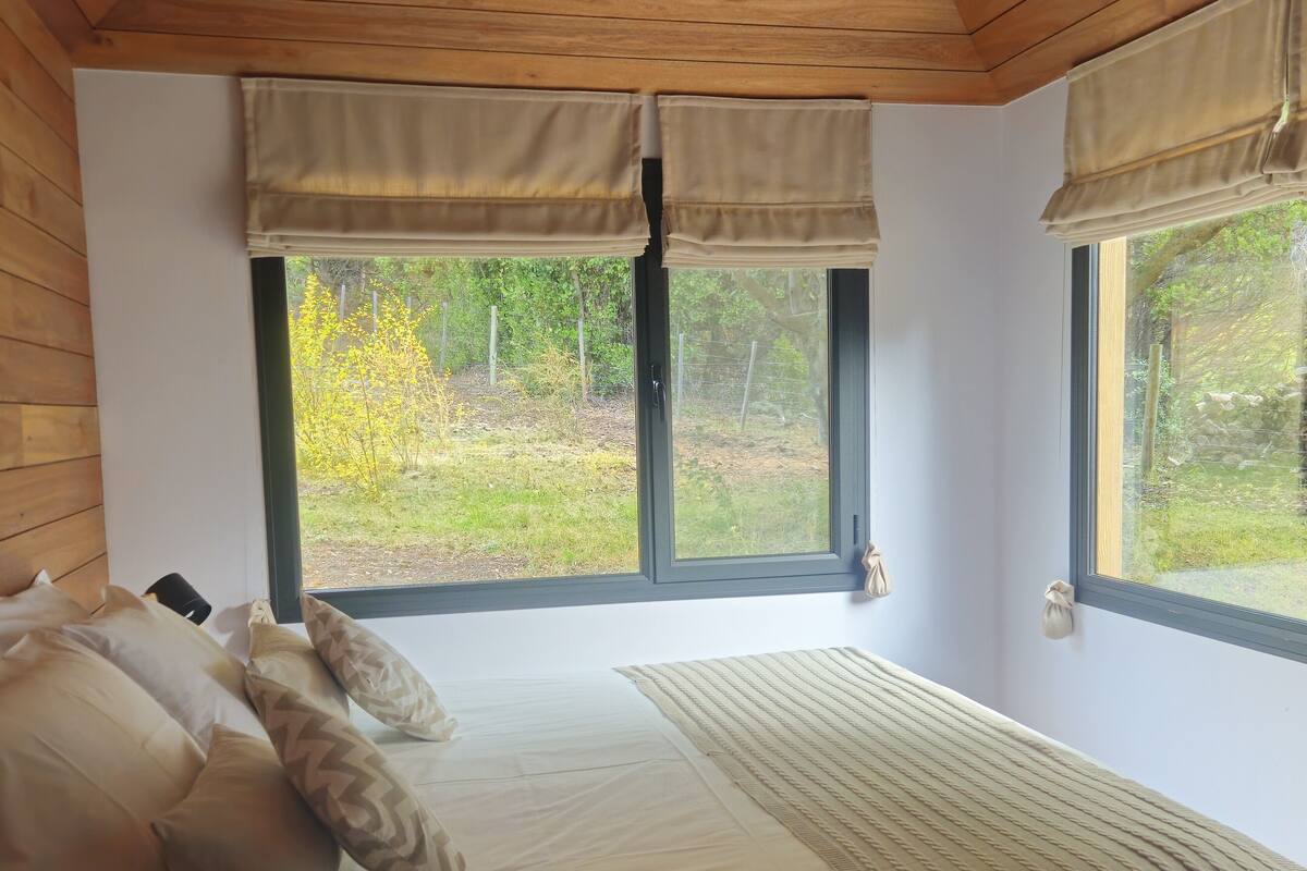 A bedroom with a bed, pillows, a bedside table, and two windows with curtains.