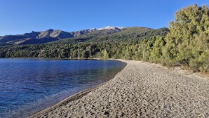 A clear view of a lake with a pebble beach and a forested mountain range in the background.