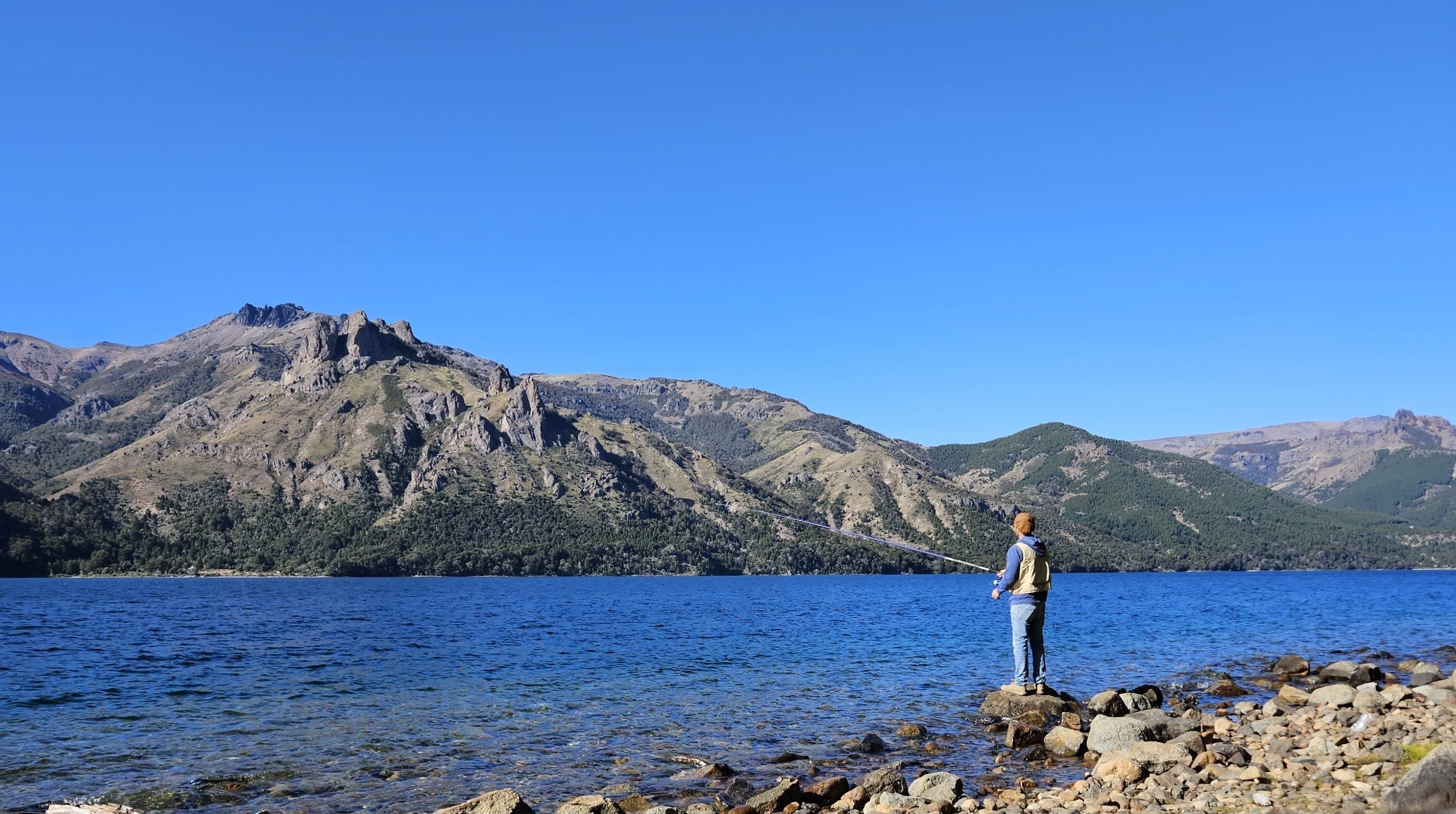 A person fishing by a rocky shoreline with mountains in the background.