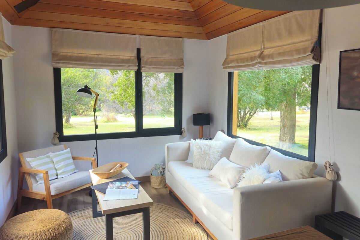 A cozy room with a white sofa, a wooden chair, a small table with a lamp, and large windows offering a view of greenery outside.