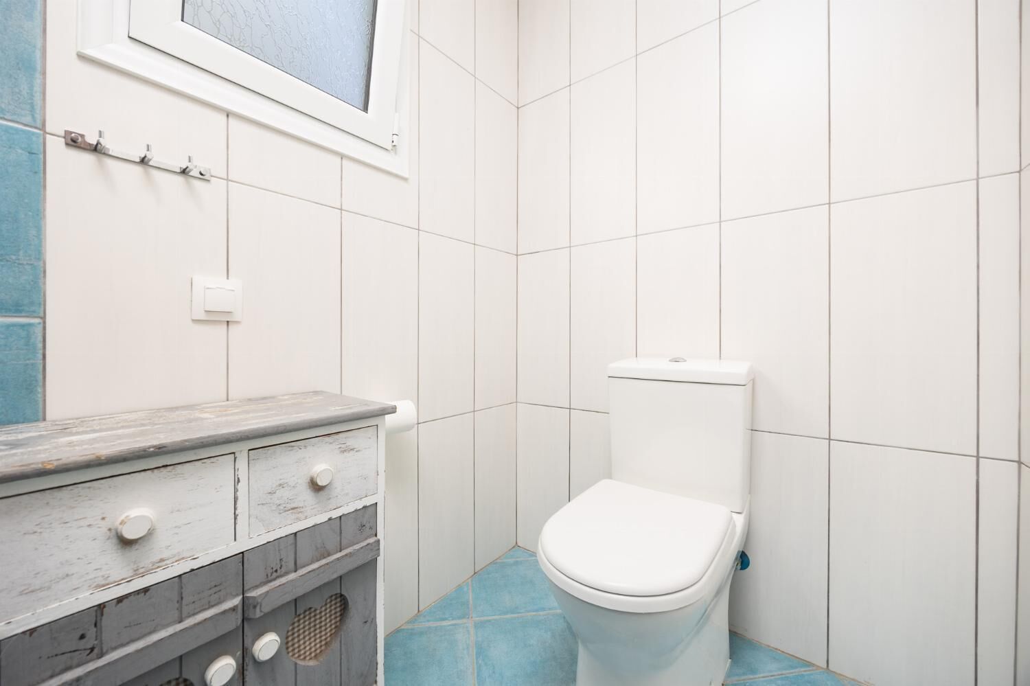A bathroom with a toilet, a wooden vanity with drawers, a window, and white tiled walls.