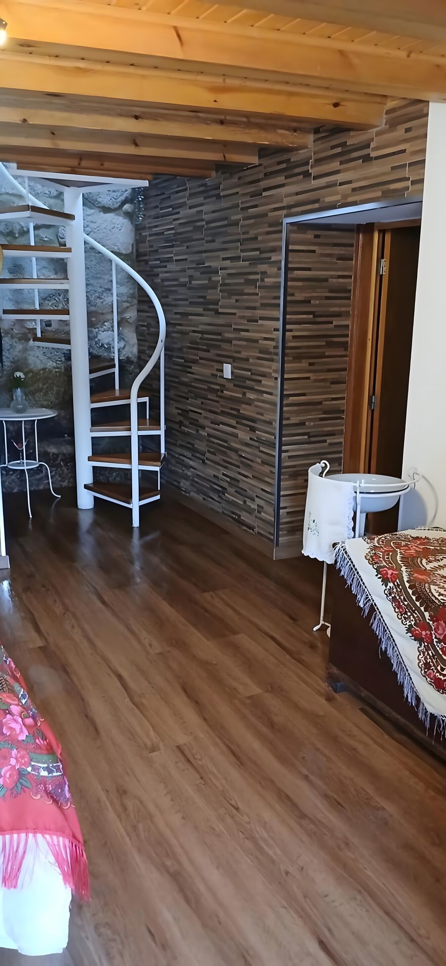 Ein Zimmer mit Wendeltreppe, einer Steinsäule, Holzdielenboden, einem Bett mit gemustertem Überwurf und einem weißen Waschbecken.