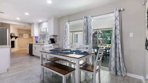 Une cuisine moderne avec des meubles blancs, une table à manger agrémentée de sets de table bleus, et une vue sur un jardin grâce à de grandes fenêtres.