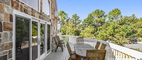 A wooden deck with a dining table and chairs, a white railing, and a stone building with glass doors.