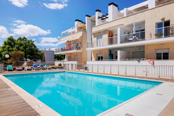 A swimming pool with lounge chairs, a wooden deck, and a multi-story building with balconies.