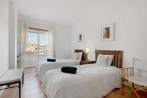 A hotel room with two beds, a bedside table, a chair, a window with curtains, and a view of buildings outside.