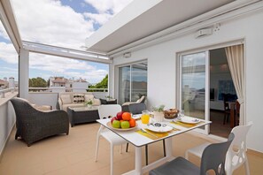 A balcony with wicker furniture, a dining table with fruit and a glass of juice, and a view of buildings and trees.
