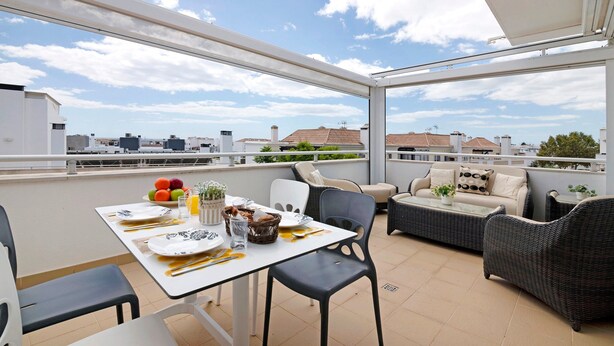 Un balcón con mesa lista para comer, mobiliario de mimbre y vista panorámica de la ciudad.