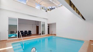 Una piscina interior con pared de vidrio, zona de estar y vista al cielo.
