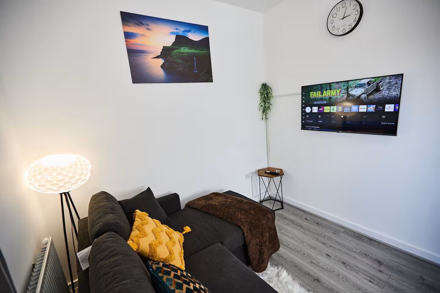 Ein modernes Wohnzimmer mit einem schwarzen Sofa, einer Lampe, einem Wand-Fernseher und einem Gemälde einer Abendlandschaft.