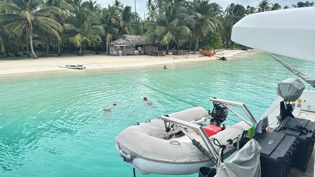 Een boot met motor en reddingsboei, een zandstrand en palmbomen op de achtergrond.