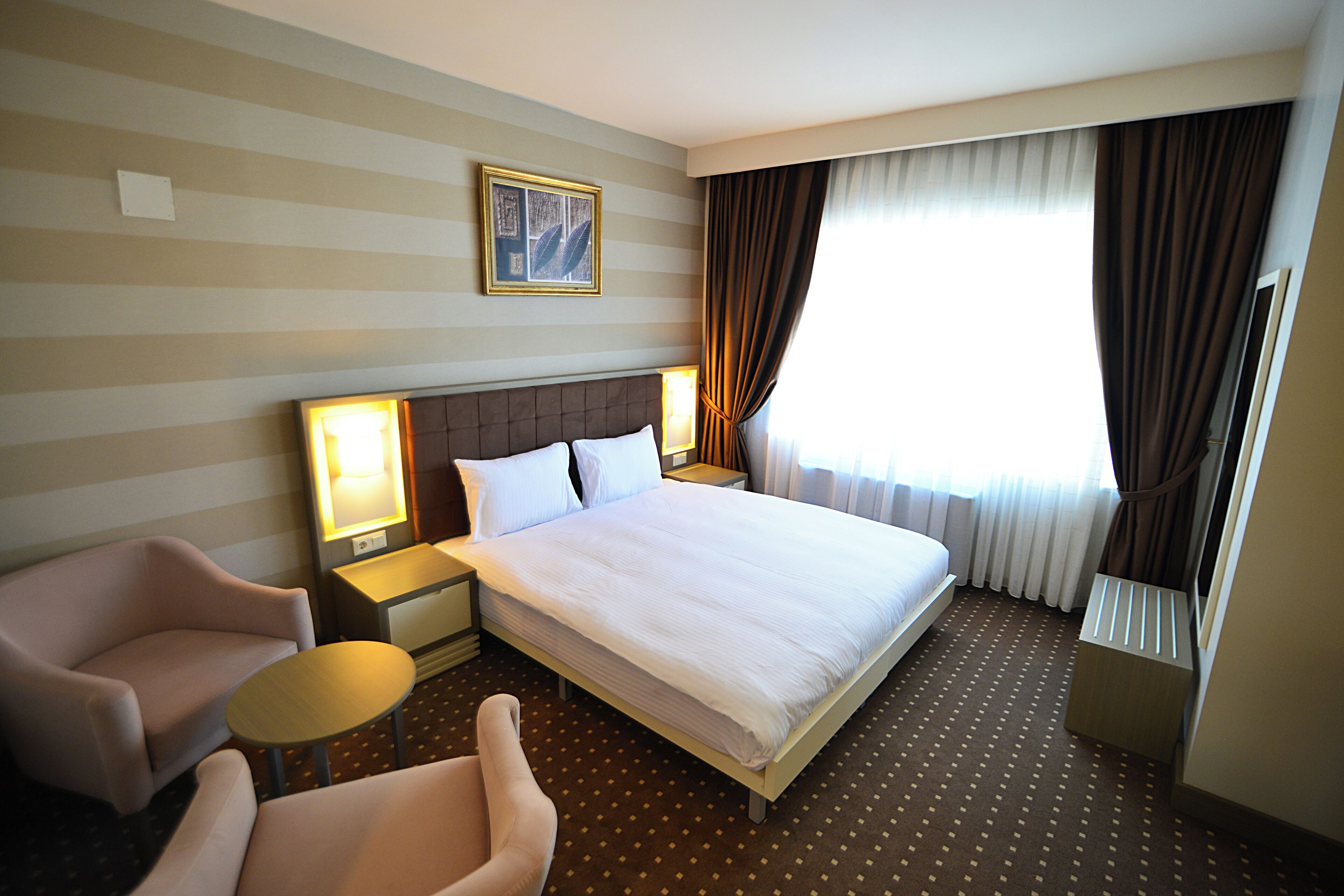 Habitación de hotel con una cama grande, dos mesitas de noche con lámparas, una silla y un ventanal con cortinas.