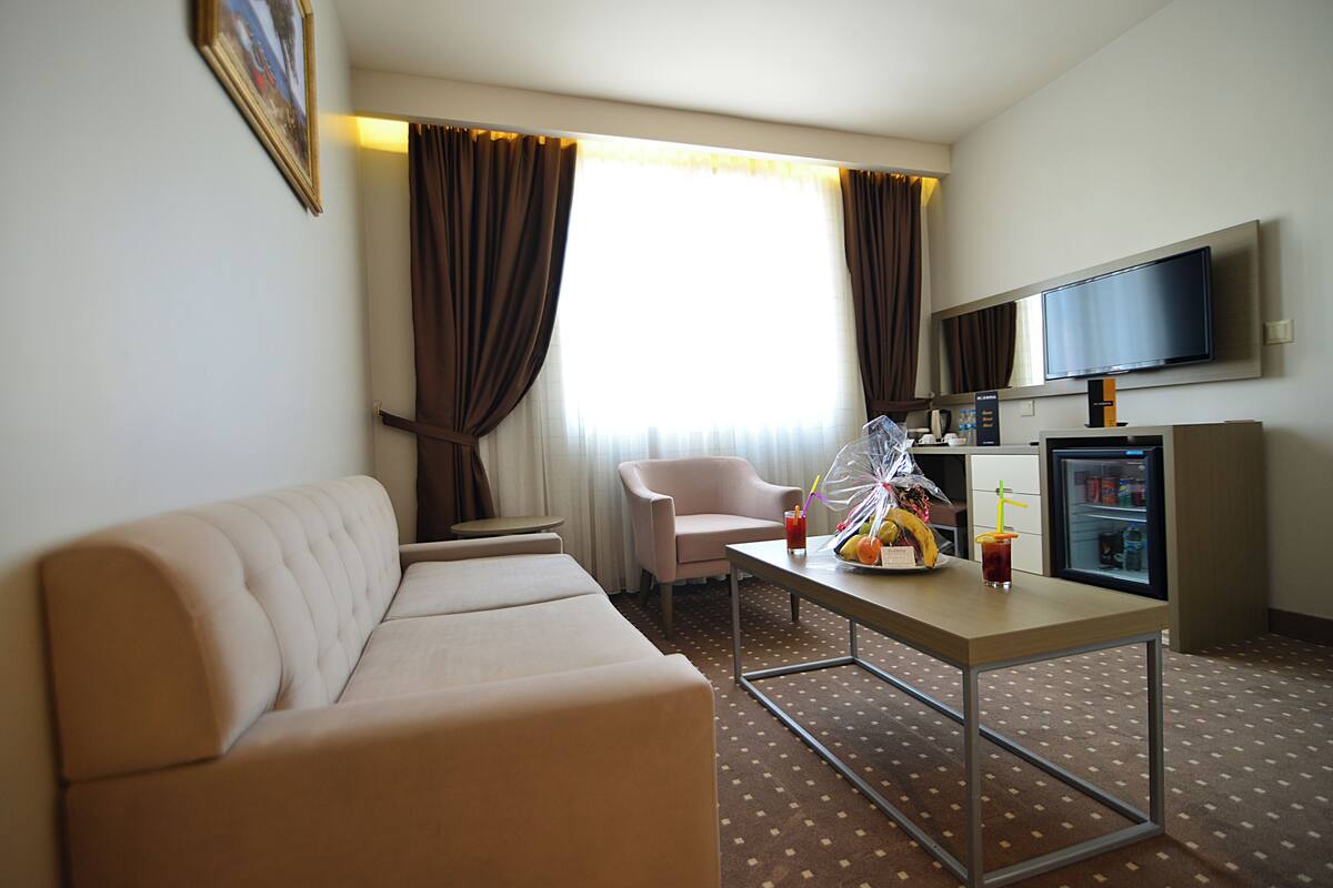 A hotel room with a sofa, a chair, a table with a drink, a TV, and a mini-fridge.
