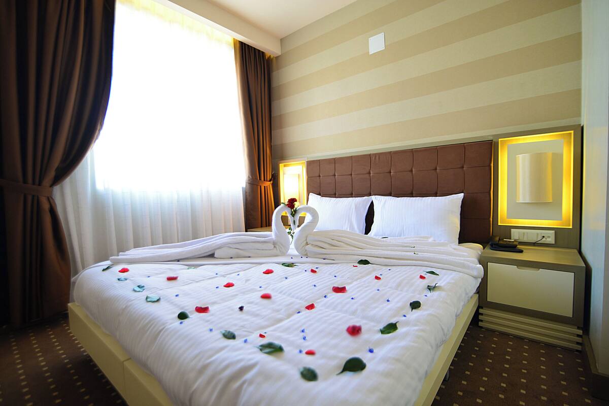 A hotel room with a neatly made bed, a bedside table with a lamp, and a window with curtains.