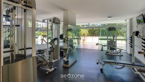 A gym with various exercise machines, a mirrored wall, and large windows offering a view of greenery outside. - Apt With River View and Balcony Pontal Bcl402 (Ilhéus)