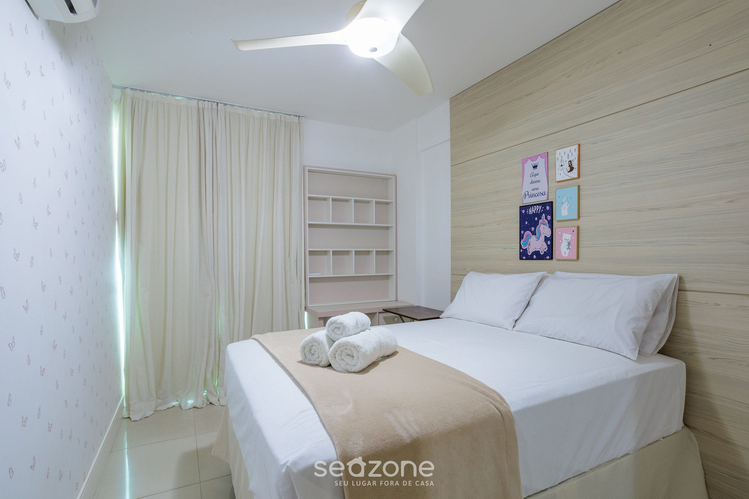 Une chambre d’hôtel comprenant un lit, une table de chevet, une étagère fixée au mur et un ventilateur de plafond.