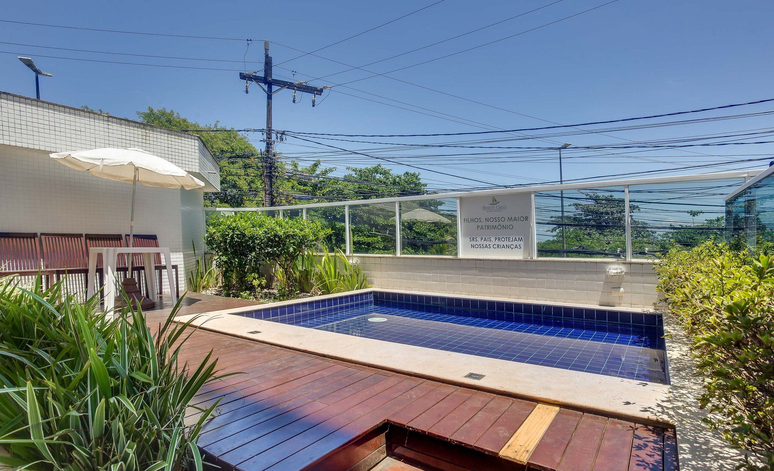 Piscina com deck revestido, cercada por vegetação e área de estar coberta.