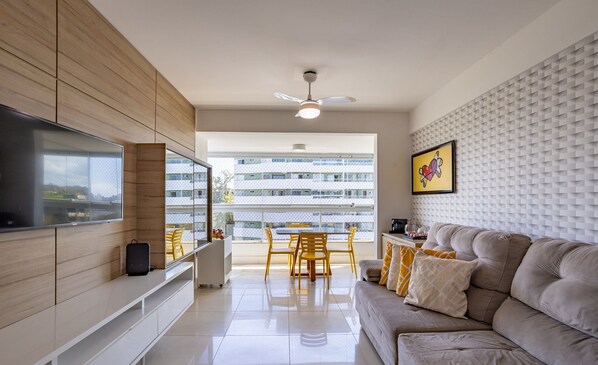 Un salon moderne doté d’un téléviseur à écran plat, d’un canapé, d’un coin repas avec des chaises et d’un balcon offrant une vue sur les bâtiments.