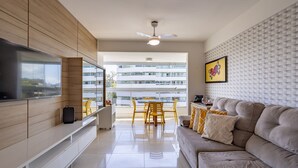 Egy modern nappali, melyben található egy síkképernyős televízió, egy kanapé, egy étkezősarok székekkel, valamint egy erkély, ahonnan kilátás nyílik az épületekre.