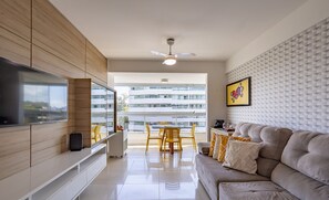 Una sala de estar moderna con televisor de pantalla plana, sofá, comedor con sillas y balcón con vistas a edificios.