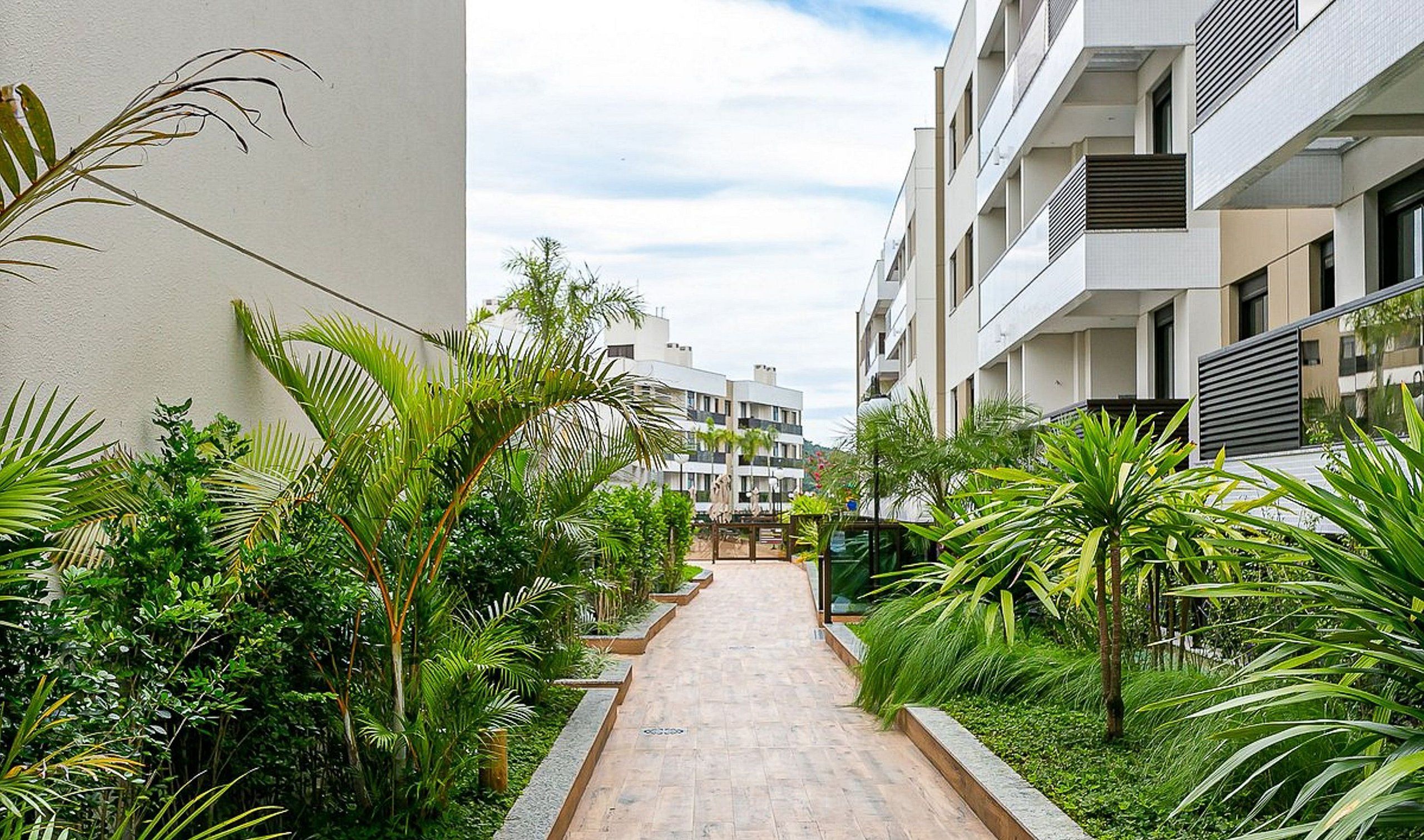 A narrow pathway flanked by lush greenery and modern buildings with balconies.