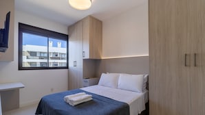 A modern bedroom with a bed, a nightstand, and a wardrobe. There is a window with a view of buildings outside. - Apart Near the Beach in Cond. w/ Pool Adc0205 (Florianópolis)