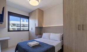 Quarto moderno com cama, mesa de cabeceira e guarda-roupa. Há uma janela com vista para edifícios.