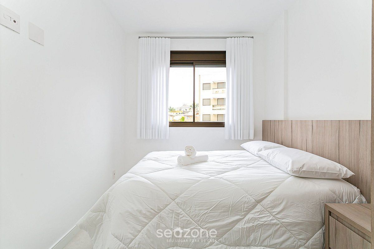 Quarto minimalista com uma cama grande, cabeceira de madeira e janela com cortinas brancas.