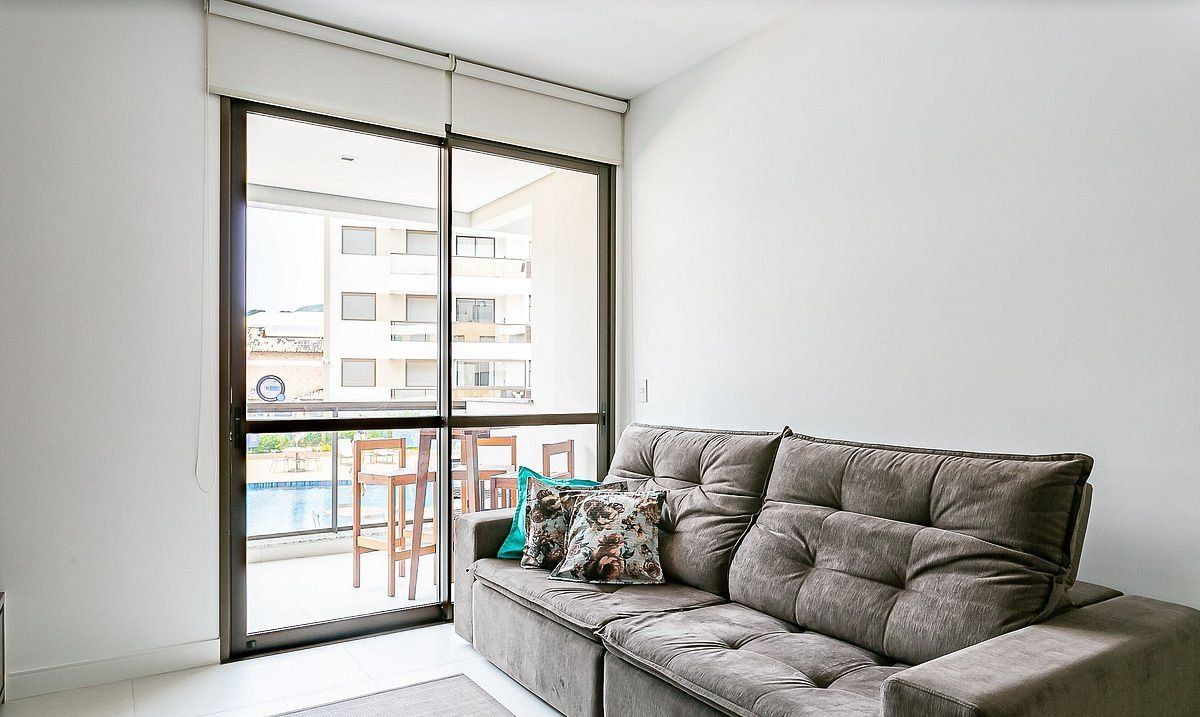 Una sala de estar moderna con un sofá gris, un ventanal corredero que da a un balcón y vistas de un edificio y una piscina.