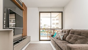 A modern living room with a flat-screen TV, a sofa, and a sliding glass door leading to a balcony with a pool. - Apartments With Pools 200m From the sea Floripa-sc ADC (Florianópolis)