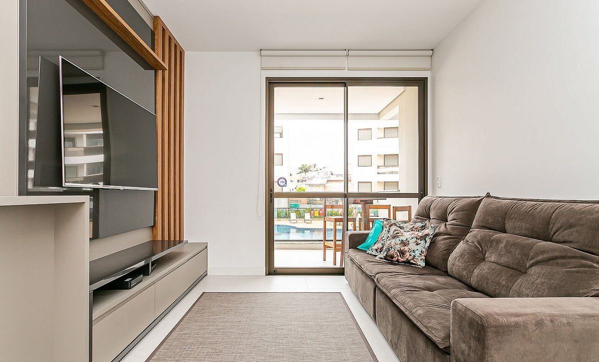 A modern living room with a flat-screen TV, a sofa, and a sliding glass door leading to a balcony with a pool.