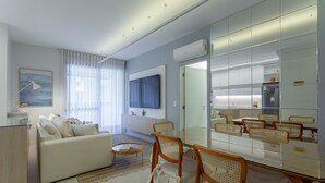 A modern living room with a sofa, coffee table, and dining area with chairs. There is a flat-screen TV mounted on the wall, a large mirror, and a refrigerator. - Apartments With Pools 200m From the sea Floripa-sc ADC (Florianópolis)