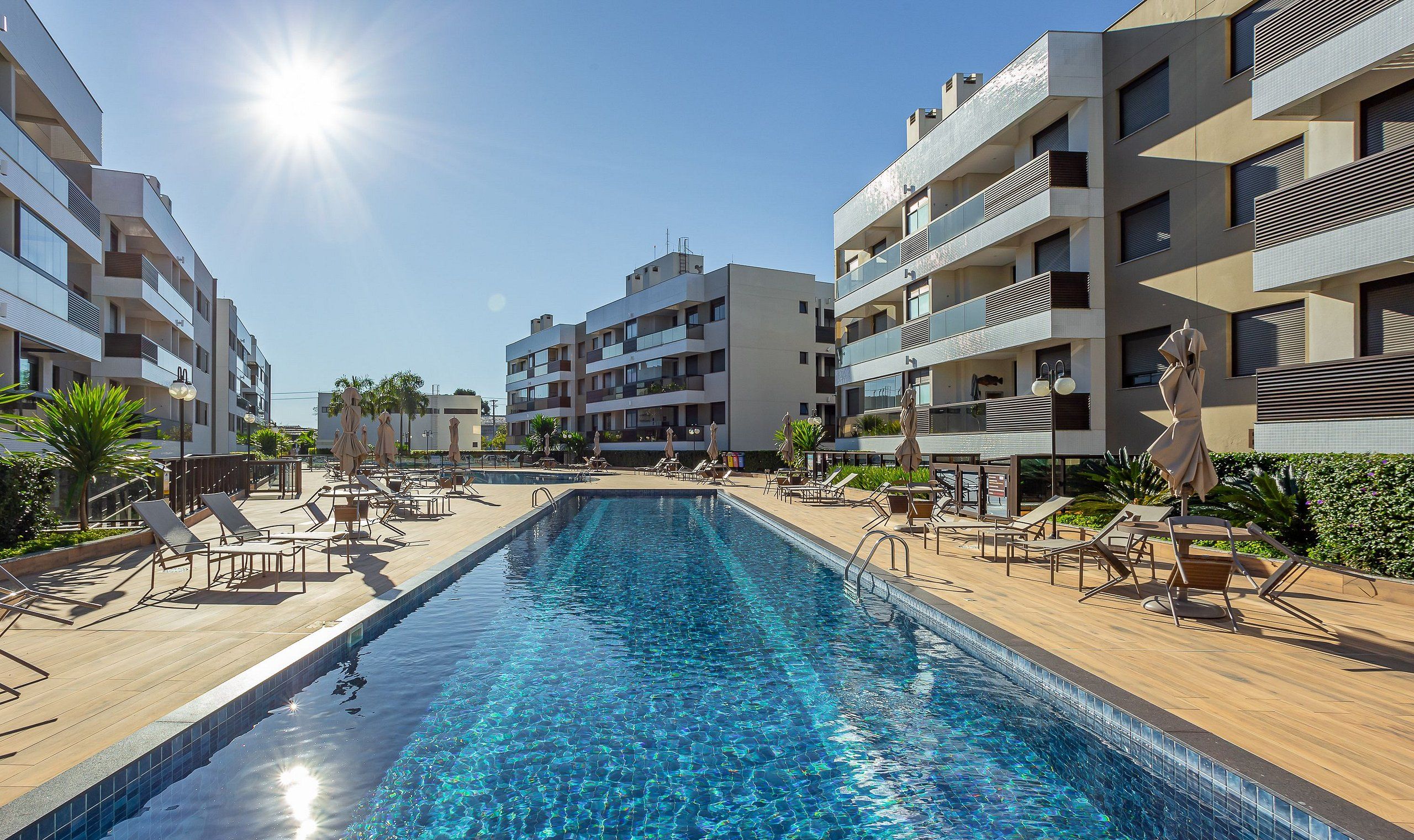 A swimming pool surrounded by sun loungers and umbrellas, with modern buildings in the background.