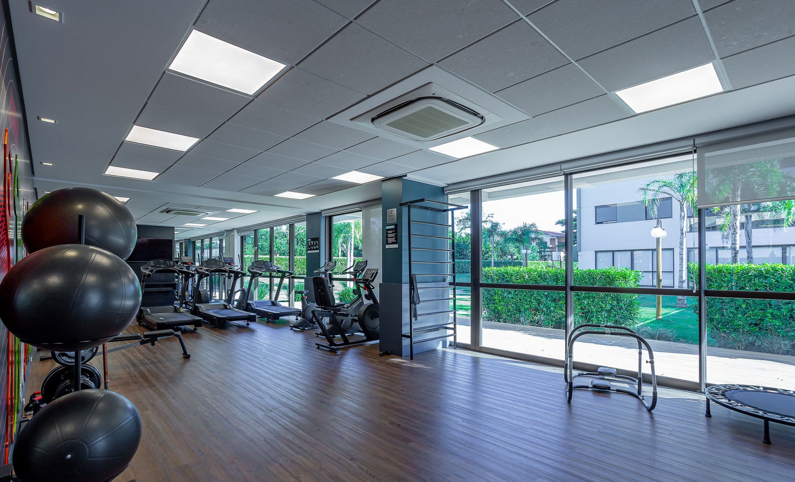 Un gimnasio amplio con cintas de correr, elípticas y pelotas de ejercicio. Amplias ventanas ofrecen vistas a zonas verdes y un edificio exterior.