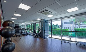 Een ruime fitnessruimte met loopbanden, elliptische trainers en fitnessballen. Grote ramen bieden uitzicht op groen en een gebouw buiten.