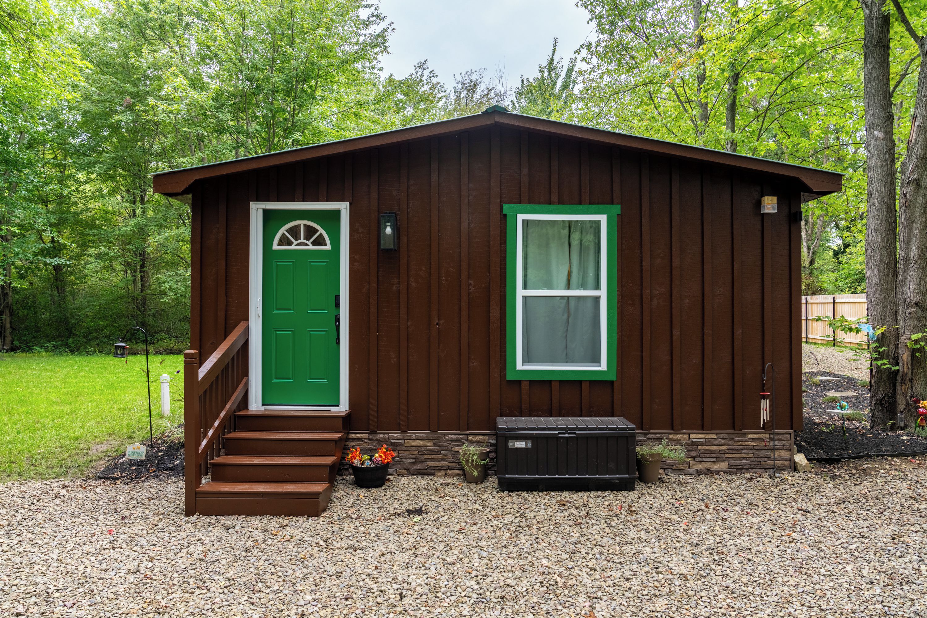 A small wooden cabin with a green door and window, a set of stairs leading to the entrance, and a black trash bin beside it.