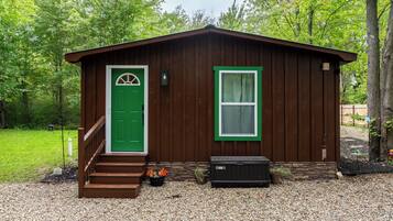 A small wooden cabin with a green door and window, a set of stairs leading to the entrance, and a black trash bin beside it.
