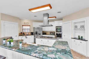 Une cuisine moderne avec des meubles blancs, un îlot central, des appareils en acier inoxydable et un pare-feu orné d’un motif en pierre naturelle.