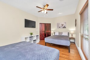 Quarto com duas camas, ventoinha no teto, televisão, porta de madeira e janela com persianas.