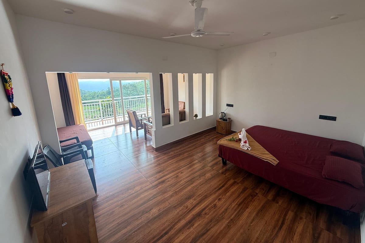 A spacious room with a wooden floor, a red sofa, a dining table with chairs, and a balcony with a view of greenery.